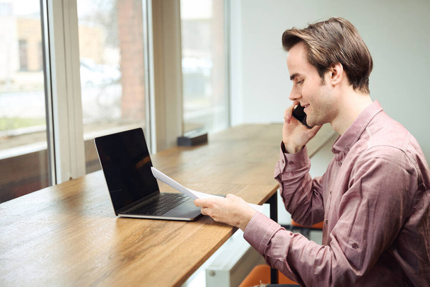 スマートフォンで話している間にノートパソコンを見て文書を見て座っている笑顔のオフィスの従業員の側面図 - 写真・画像