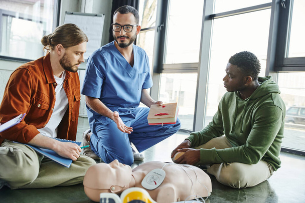 smiling healthcare worker in eyeglasses and uniform holding wound care simulator and pointing at CPR manikin near multicultural participants of medical seminar, effective life-saving skills concept - Photo, Image