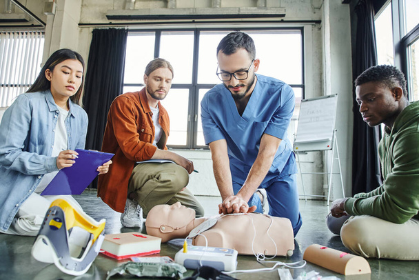 νεαρή Ασιάτισσα με πρόχειρο που κοιτάζει τραυματιοφορέα να κάνει μαλάξεις στο στήθος στο ανδρείκελο CPR κοντά σε ιατρικό εξοπλισμό και πολυεθνικούς συμμετέχοντες σε σεμινάριο πρώτων βοηθειών στην αίθουσα εκπαίδευσης - Φωτογραφία, εικόνα