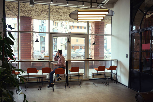 Радостный парень с кучей документов в руке, сидящий за столом у окна во время разговора по телефону - Фото, изображение