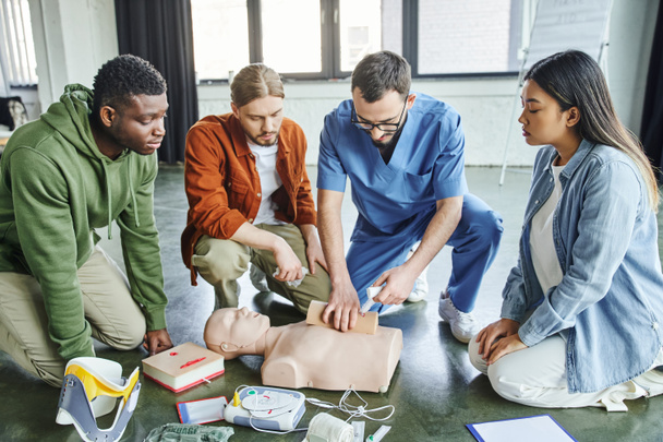 CPRマニキン、除細動器、医療機器の近くの多文化チームに救命スキルを示しながら、包帯でシミュレータに傷をタンピング医療従事者、緊急時対応コンセプト - 写真・画像