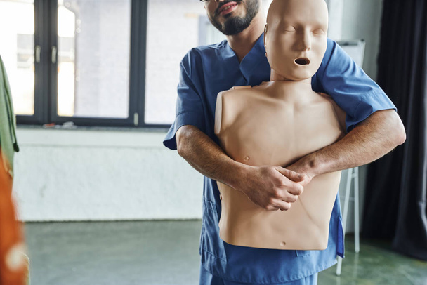 first aid training seminar, partial view of bearded medical instructor in uniform practicing life-saving techniques in case of chocking on CPR manikin, emergency situations preparedness concept - Photo, Image