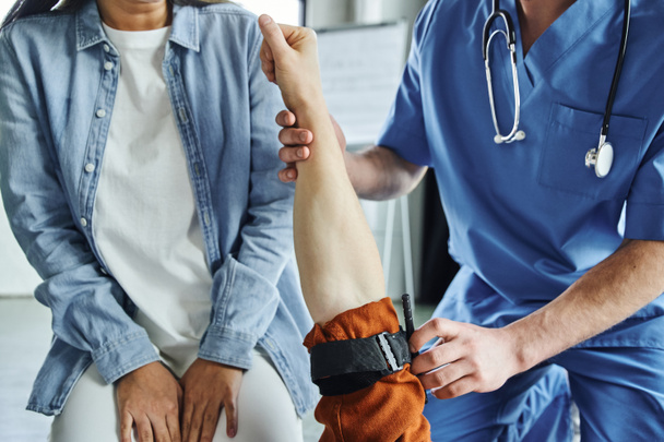 Ausgeschnittene Ansicht eines professionellen Sanitäters, der Kompressionstour am Arm eines Mannes in der Nähe einer jungen Frau während eines Erste-Hilfe-Seminars im Schulungsraum anwendet, Reaktionskonzept für Notfallsituationen - Foto, Bild