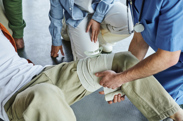 μερική άποψη του επαγγελματία τραυματιοφορέα που εφαρμόζει συμπιεστικό επίδεσμο στο πόδι του ανθρώπου κοντά σε πολυεθνική ομάδα κατά τη διάρκεια σεμιναρίου κατάρτισης πρώτων βοηθειών, έννοια τεχνικών πρόληψης αιμορραγίας - Φωτογραφία, εικόνα