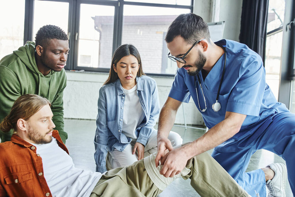 professional healthcare worker applying compression bandage on leg of man near multiethnic students during first aid seminar in training room, importance of emergency preparedness concept - Photo, Image