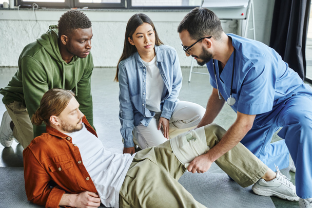 νοσηλευτής που εφαρμόζει συμπιεστικό επίδεσμο στο πόδι του ανθρώπου κοντά σε διαφυλετικούς συμμετέχοντες κατά τη διάρκεια σεμιναρίου πρώτων βοηθειών στην αίθουσα εκπαίδευσης, έννοια τεχνικών πρόληψης αιμορραγιών - Φωτογραφία, εικόνα