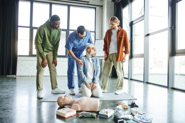 praktická první pomoc učení, mladí mezirasoví muži při pohledu na zdravotnického pracovníka obvazování hlavy asijské ženy v blízkosti lékařského vybavení a CPR figuríny ve výcvikové místnosti, koncept nouzové reakce - Fotografie, Obrázek