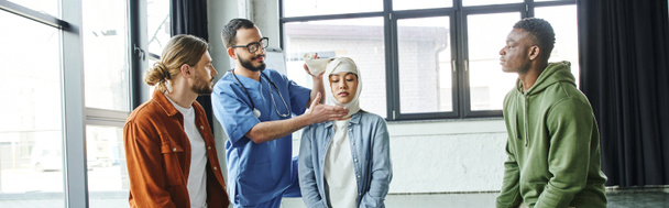 lächelnder Sanitäter mit Brille, der den Kopf einer jungen asiatischen Frau in der Nähe multiethnischer Männer bandagiert, während er lebensrettende Fähigkeiten auf einem Erste-Hilfe-Seminar zeigt, Bedeutung des Notfallvorsorgekonzeptes, Banner - Foto, Bild