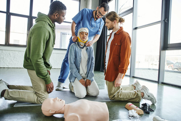 Erste-Hilfe-Training, interrassische Männer schauen auf medizinische Ausbilder, die asiatische Frau mit bandagiertem Kopf in der Nähe von CPR-Schaufensterpuppen und medizinischem Gerät mit Nackenstütze ausstatten, Reaktionskonzept für Notfallsituationen - Foto, Bild