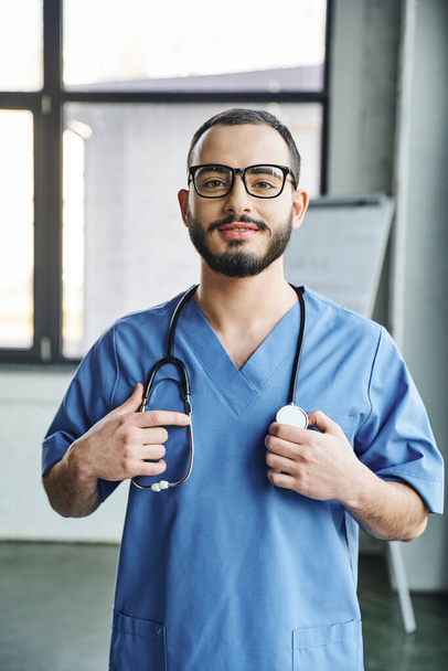 vidám szakállas egészségügyi dolgozó ragyogó mosollyal áll kék egyenruhában és megérinti sztetoszkóppal a nyakon a klinikán, elsősegély-tréning szeminárium és sürgősségi felkészültség koncepció - Fotó, kép