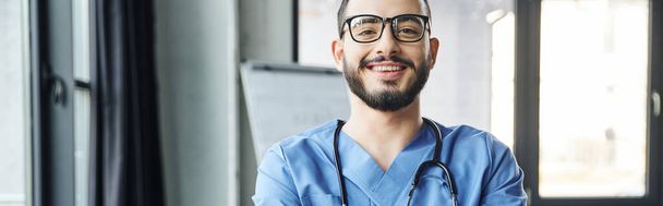 portrait of young and joyful healthcare worker with beard, eyeglasses and stethoscope looking at camera in modern clinic, first aid training seminar and emergency preparedness concept, banner - Photo, Image