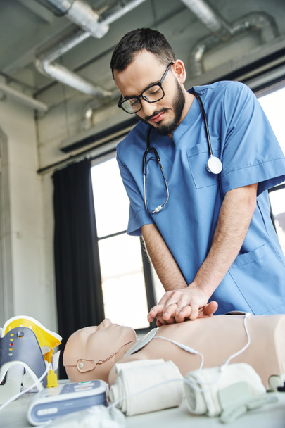 bärtiger professioneller Rettungssanitäter mit Brille und blauer Uniform, der Brustkompressionen an CPR-Schaufensterpuppe in der Nähe von Defibrillator und Druckverband übt, Konzept zur Entwicklung kritischer Fähigkeiten - Foto, Bild