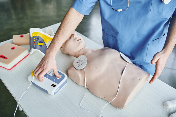 Tıbbi eğitmenin otomatikleştirilmiş defibrilatörünün CPR manikin, yara bakım simülatörleri ve boyunluk, ilk yardım eğitimi ve kritik beceri geliştirme konsepti yakınlarındaki kısmi görüntüsü - Fotoğraf, Görsel
