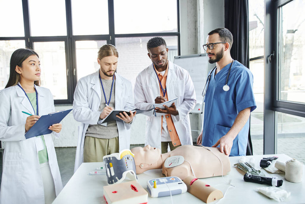 first aid seminar, diverse group of multiethnic students in white coats writing near paramedic, CPR manikin, defibrillator and medical equipment in training room, energy situations response concept - Photo, Image