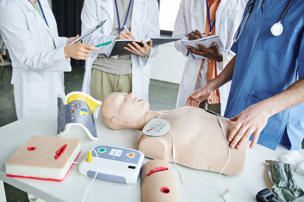 cropped view of woman in white coat pointing at CPR manikin and defibrillator near paramedic and multiethnic students writing in notebooks, life-saving skills hands-on learning concept - Photo, Image
