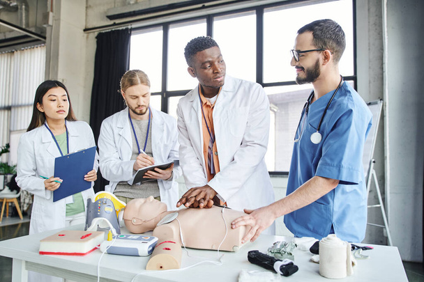 θετική ιατρική εκπαιδευτής κοιτάζοντας την αφρικανική Αμερικανός άνθρωπος εξάσκηση μαλάξεις στο στήθος σε CPR manikin κοντά σε ιατρικό εξοπλισμό και πολυεθνική φοιτητές, έκτακτης ανάγκης κατάσταση έννοια απόκρισης - Φωτογραφία, εικόνα