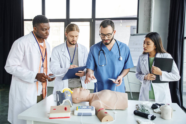 resuscytacja serca, instruktor medyczny trzymający defibrylatory powyżej manekina resuscytacyjnego w pobliżu młodych wielonarodowych studentów w białych fartuchach, koncepcja reakcji w sytuacjach awaryjnych - Zdjęcie, obraz