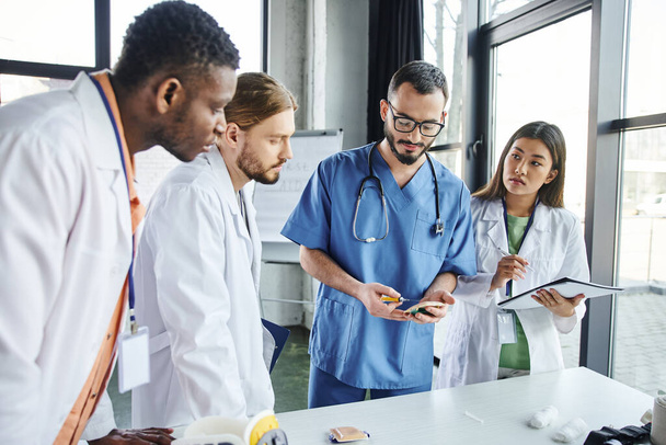 研修室での医療セミナーでは白衣を着た多文化の学生の多様なグループの近くに注射パッド付きのプロの救急医療の練習,スキル開発の概念 - 写真・画像
