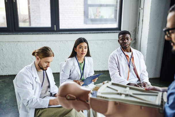 medical training, diverse group of multiethnic students in white coats looking at paramedic standing with CPR manikin on blurred foreground, acquiring and practicing life-saving skills concept - Photo, Image