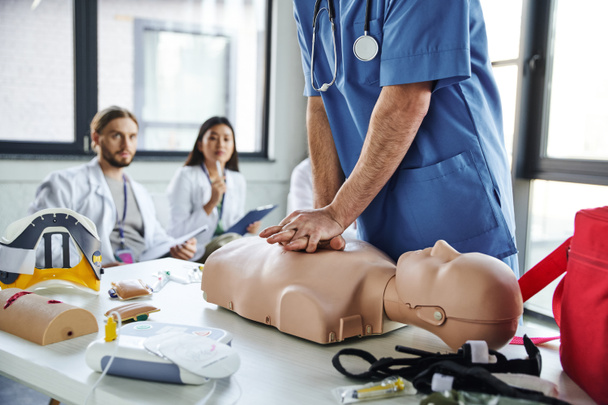 επαγγελματίας τραυματιοφορέας με μπλε στολή που κάνει μαλάξεις στο στήθος στο ανδρείκελο CPR κοντά σε ιατρικό εξοπλισμό και νέους πολυεθνικούς φοιτητές σε θολή φόντο, εξάσκηση στην έννοια της διάσωσης δεξιοτήτων - Φωτογραφία, εικόνα