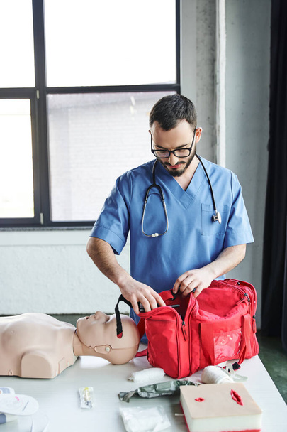 young medical instructor in eyeglasses and blue uniform preparing training room for first aid seminar and opening red bag near CPR manikin, life-saving skills development concept - Photo, Image