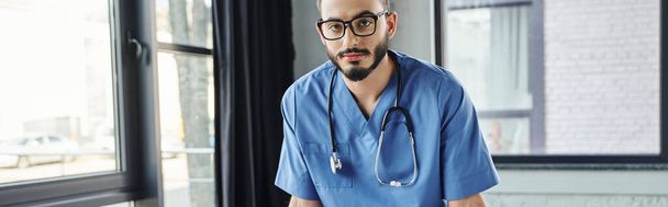 serious and bearded healthcare worker in eyeglasses and blue uniform looking at camera in training room before first aid seminar, life-saving skills development concept, banner - Photo, Image