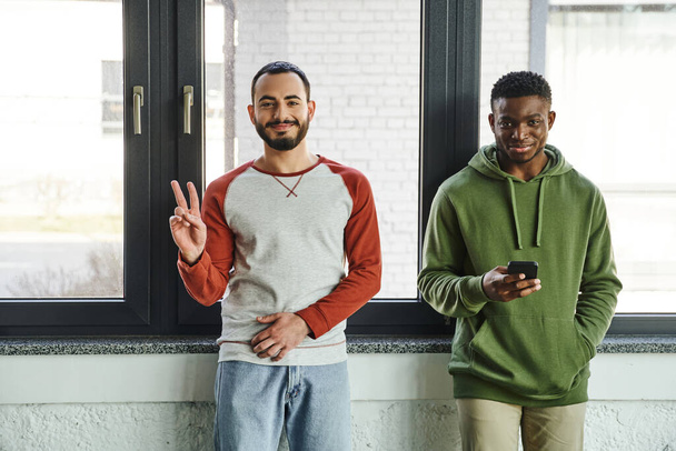 young interracial business colleagues in stylish casual clothes looking at camera in modern office, cheerful bearded man showing victory sign near african american man networking on mobile phone - 写真・画像
