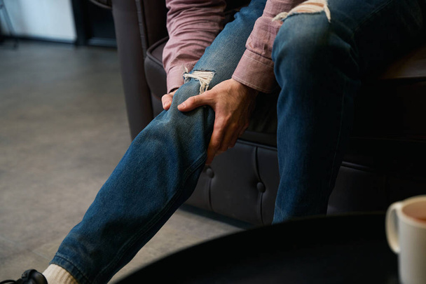 Foto cortada do homem tocando sua rótula enquanto sentado no sofá de couro - Foto, Imagem