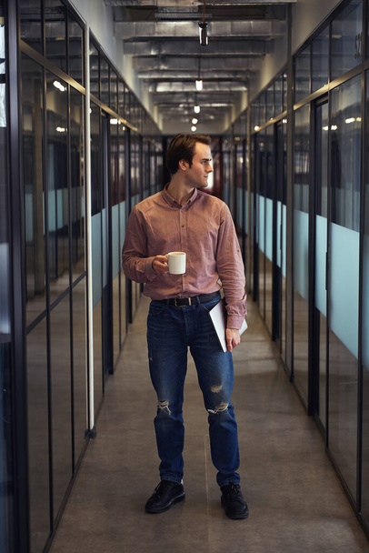 Retrato de larga duración del trabajador corporativo tranquilo con portátil y taza en las manos de pie en el pasillo de la oficina vacía - Foto, Imagen