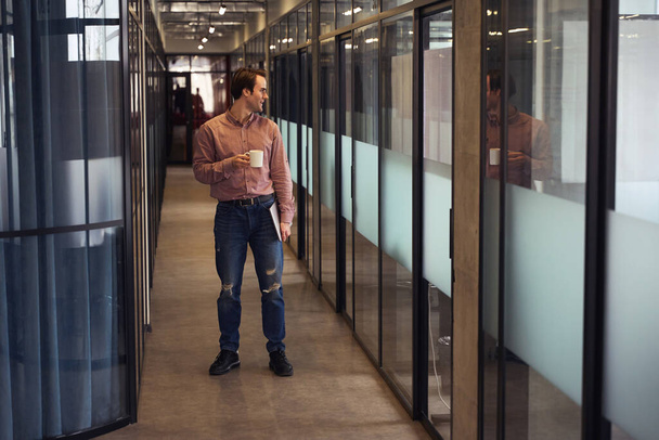Pleased man with computer and cup in hands looking at his reflection on glass wall in empty office corridor - 写真・画像