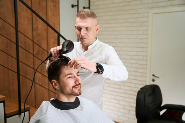 Master puts the hair to the client with a comb and hairdryer, modern interior in the barbershop - Foto, afbeelding