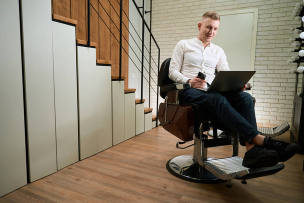 Man in a light shirt sits in a hairdressing chair, he has a laptop and a phone in his hands - Photo, image