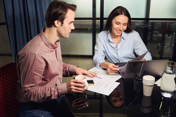 Mujer sonriente gerente de oficina sentada en el portátil junto a su asistente personal mirando el documento en sus manos - Foto, Imagen