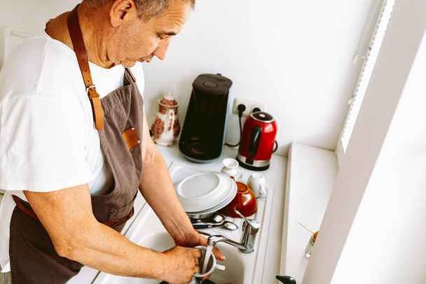 middle-aged man, in brown chefs apron, with muscular arms, washes dishes in kitchen sink, at home. Homework, help. Husband helps wife wash dishes - Foto, Imagen