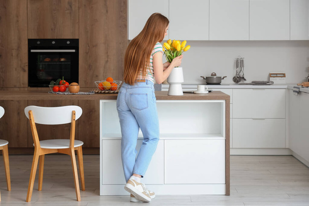 Beautiful young woman smelling blooming tulip flowers in modern kitchen - Foto, Imagen