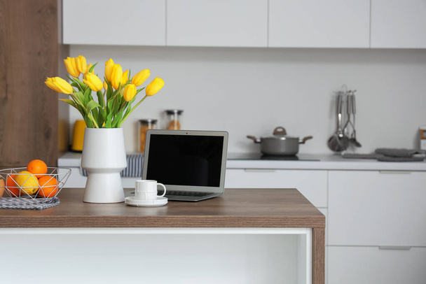 Vase with yellow tulip flowers, modern laptop, fruits and cup of coffee on wooden table in light kitchen - Фото, зображення