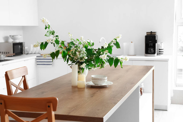 Vaas met bloeiende jasmijnbloemen, kaarsen en schalen op houten tafel in moderne keuken - Foto, afbeelding