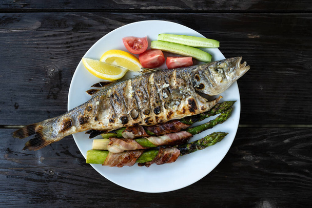 Grilled sea bass fish with lemon pieces, fresh salad and grilled asparagus with bacon on white plate. Food photography - Photo, Image