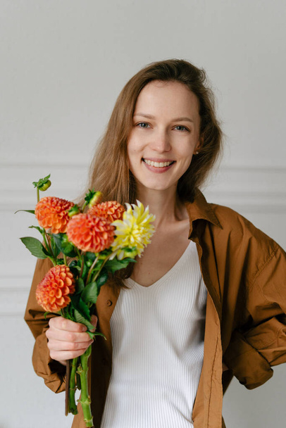 Retrato de una joven florista sonriente elegantemente vestida sosteniendo un ramo de dalias sobre un fondo blanco. Concepto idea para regalos, flores de temporada y floristería - Foto, imagen