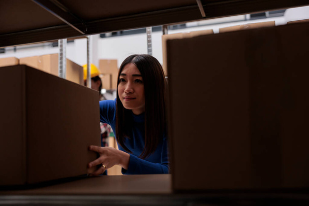 Distribution center worker taking cardboard box from rack to prepare goods for dispatching. Asian woman order picker searching carton package on shelf in industrial warehouse - Zdjęcie, obraz