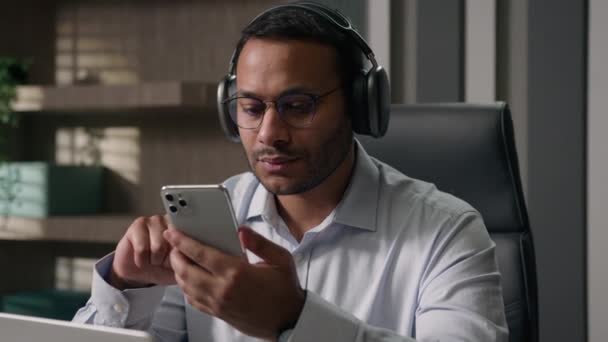 Αμερικανός επιχειρηματίας Ινδός άνθρωπος Άραβας εργαζόμενος αφεντικό στα ακουστικά ακούσετε μουσική χρησιμοποιώντας το κινητό τηλέφωνο επιλέξτε το τραγούδι σε ηχητική εφαρμογή στο τραπέζι του γραφείου ήρεμο ήχο χαλαρωτικό διάλειμμα εργασίας τέλος ήρεμο χαλαρώστε - Πλάνα, βίντεο