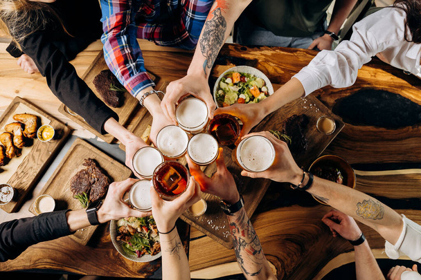 Amigos vitoreando vasos de cerveza en una mesa de madera cubierta con deliciosa comida - Vista superior de la gente teniendo una cena en el bar restaurante - Concepto de estilo de vida de alimentos y bebidas - Foto, Imagen