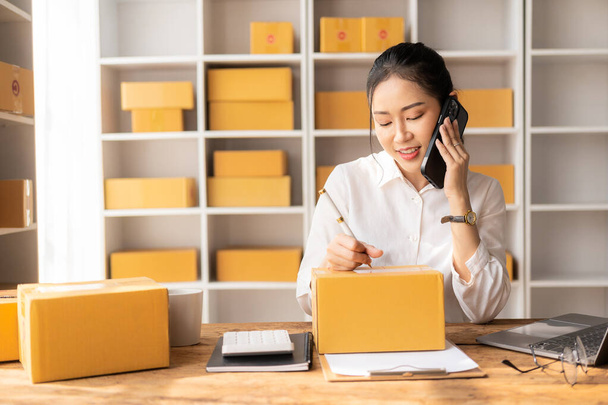 Small Business Startup, SME, Entrepreneur - Gelukkige Aziatische vrouw die smartphone of laptop gebruikt om online bestellingen te ontvangen en te verifiëren om dozen voor te bereiden en pakketten van online bestelling te leveren. - Foto, afbeelding