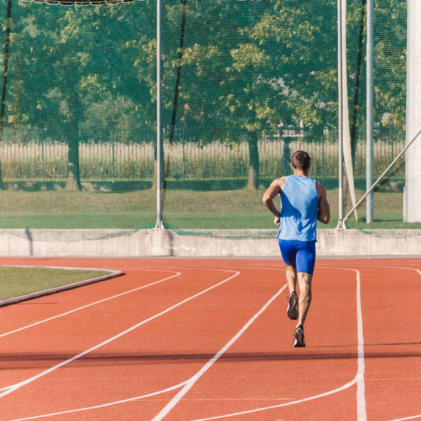 Caucasian male athlete in blue sportswear running alone along an athletic track on the stadium - 写真・画像