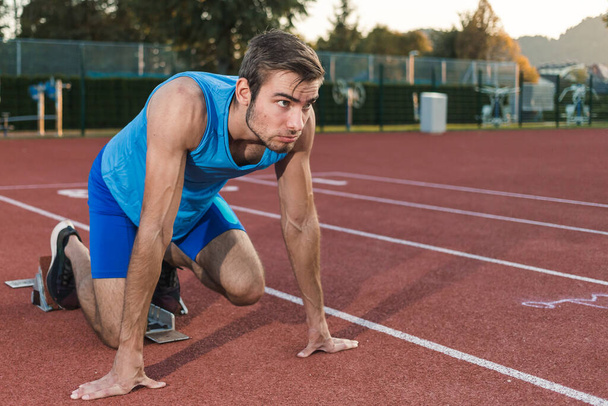 Young professional male athlete in blue sportswear taking a start position for a sprint run during outdoor training - Photo, Image