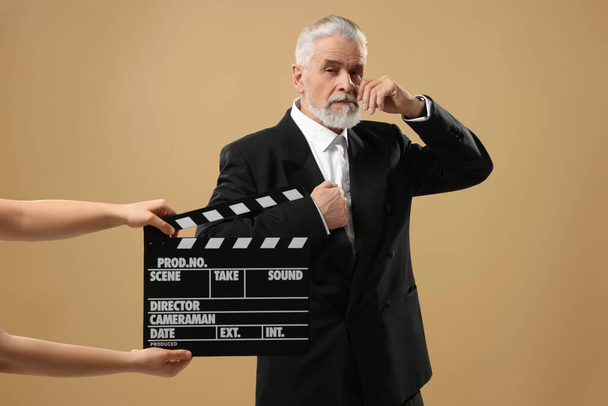 Senior actor performing role while second assistant camera holding clapperboard on beige background. Film industry - Фото, изображение
