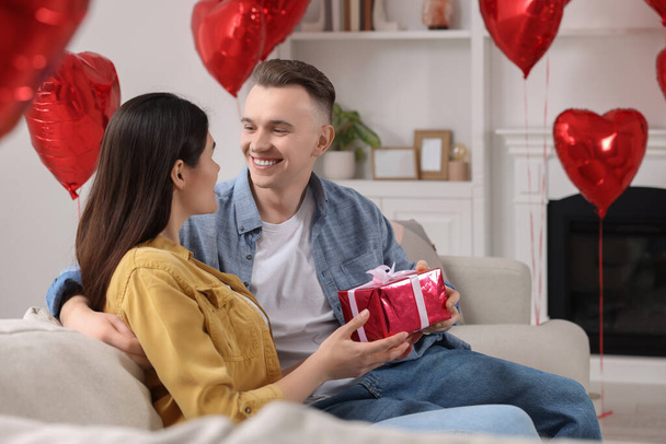 Счастливая пара празднует День Святого Валентина. Любимая женщина с подарочной коробкой в комнате, украшенной воздушными шарами в форме сердца - Фото, изображение