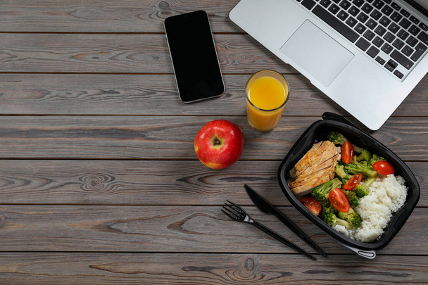 Container of tasty food, glass of juice, laptop, apple and smartphone on wooden table, flat lay with space for text. Business lunch - Fotó, kép