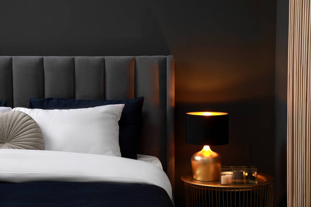 Comfortable bed with cushions, lamp and different decor on bedside table in room. Stylish interior - Foto, imagen