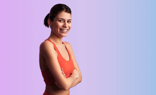 Positive young athletic female in red sports top standing with crossed hands against gradient background looking at camera - Foto, immagini